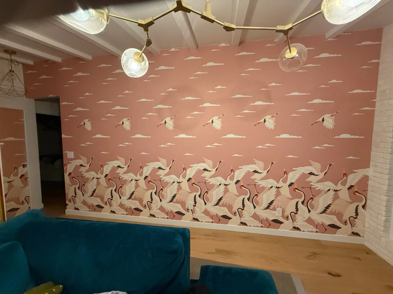 Living room with blush crane-and-cloud wallcovering, teal seating, and brass light fixtures.